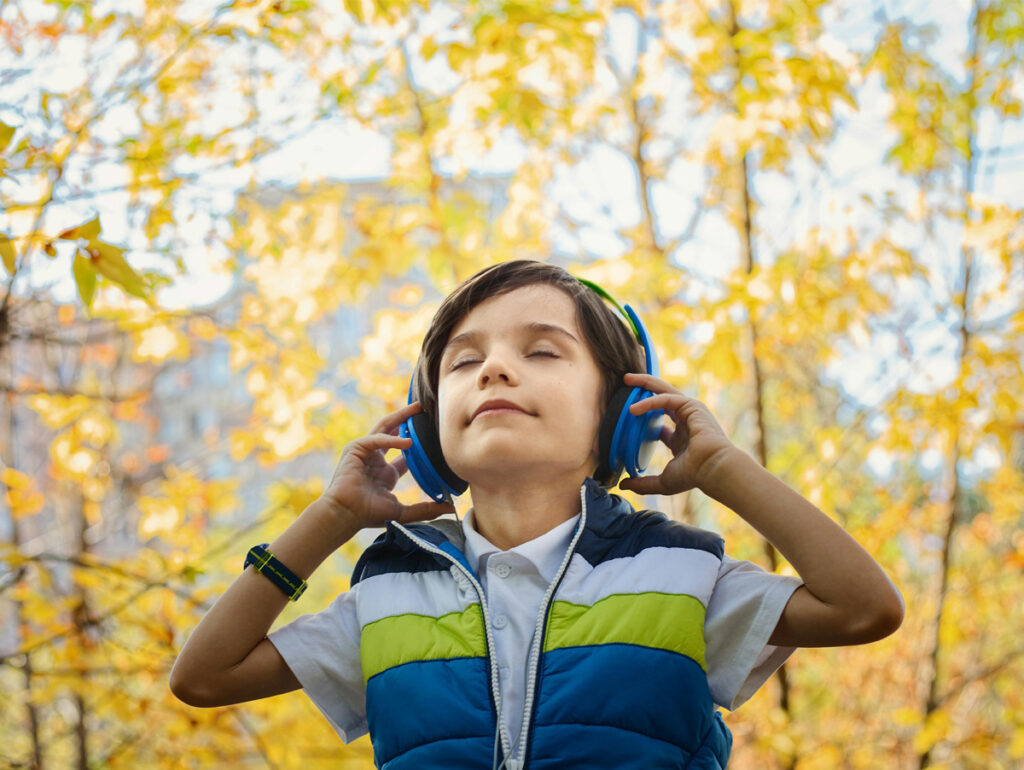 Criança feliz ouvindo música com fones de ouvido azuis em um ambiente natural com folhas amarelas ao fundo, expressando relaxamento e alegria.
	
